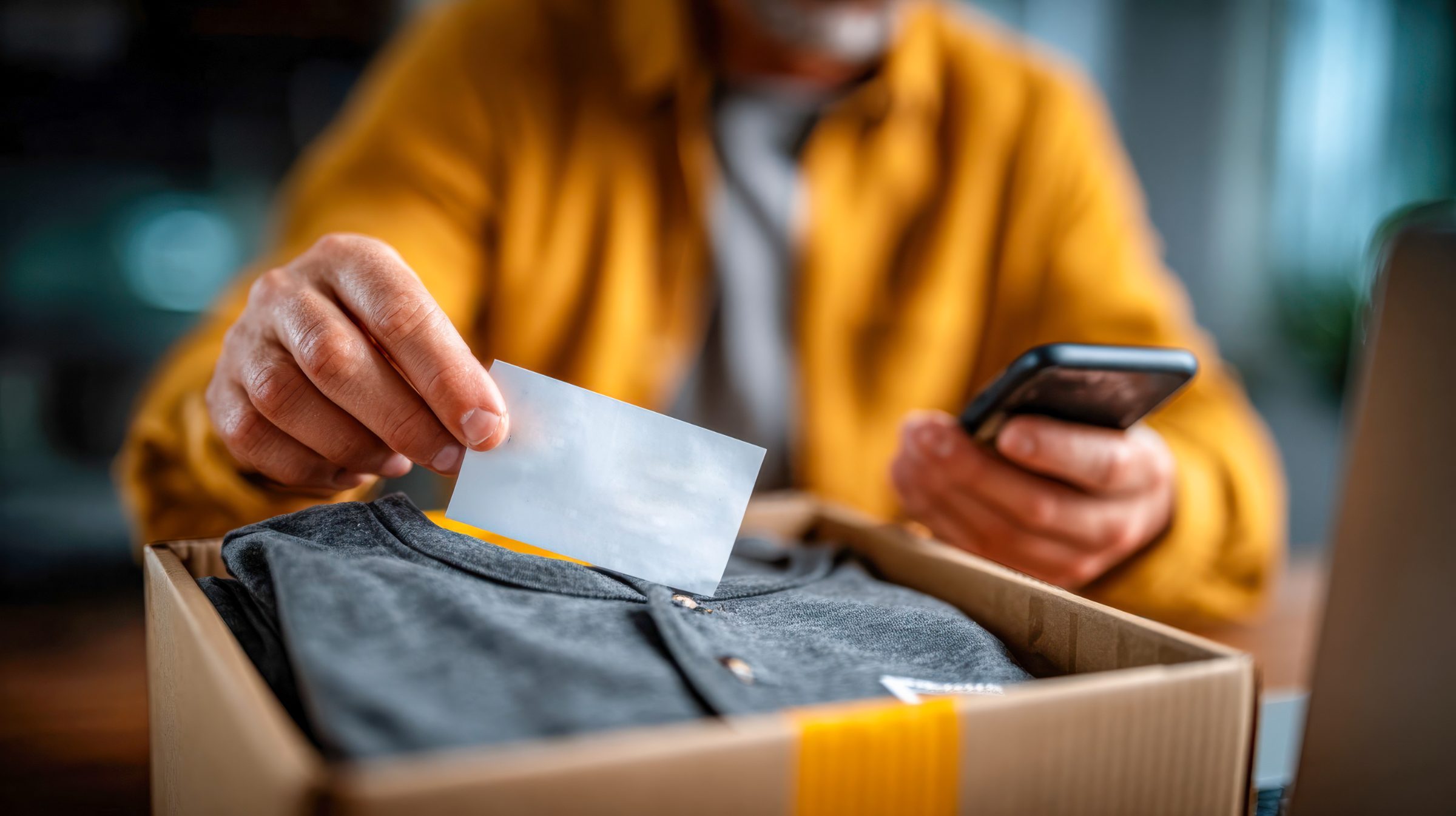 A sender tucking a note into a packed parcel, phone in hand